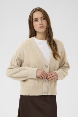 Frau mit langen Haaren, die Knöpfe eines beigen, flauschigen Cardigans über einem weißen Oberteil und einem dunkelbraunen Rock vor einfachem Hintergrund zumacht.