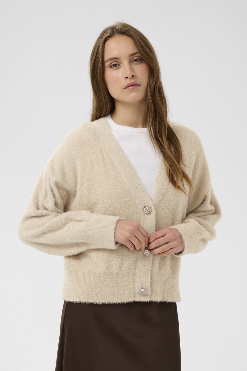 Žena s dlouhými vlasy zapíná knoflíky na béžovém huňatém svetru přes bílý top a tmavě hnědou sukni na jednobarevném pozadí.