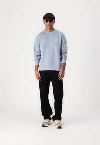 Lyseblå hettegenser med rund hals og logo-detalj, kombinert med svarte bukser og beige joggesko med hvite detaljer.