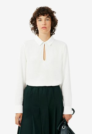 Persoon met krullend haar draagt een witte blouse met lange mouwen en sleutelgat halslijn en een donkere geplooide rok, houdt een zwarte handtas vast.