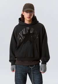 Schwarz gefütterter Kapuzenpullover mit einem strukturierten Sternendesign, lockere Passform und Kängurutasche. Kombiniert mit dunklen Jeans und grauen Fingerhandschuhen.