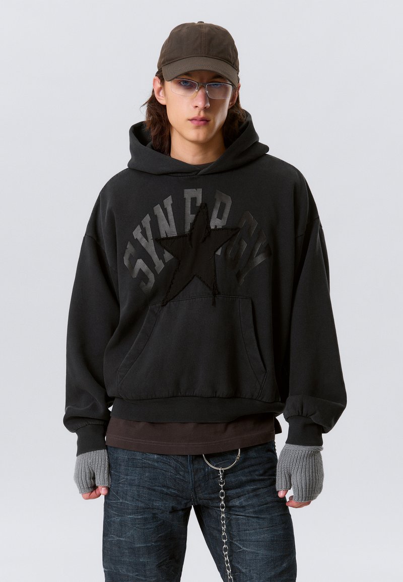 Schwarz gefütterter Kapuzenpullover mit einem strukturierten Sternendesign, lockere Passform und Kängurutasche. Kombiniert mit dunklen Jeans und grauen Fingerhandschuhen.