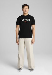 Sort t-shirt med "PORTUGAL" i hvid tekst og Puma-logo i rød. Parret med beige vidde bukser og grå sko. Ensfarvet baggrund.