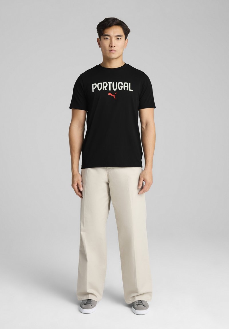 Sort t-shirt med "PORTUGAL" i hvid tekst og Puma-logo i rød. Parret med beige vidde bukser og grå sko. Ensfarvet baggrund.