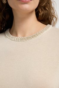 Cremfarbener Pullover mit strukturiertem Finish, ausgestattet mit einem mit Juwelen verzierten Kragen mit klaren Perlen und goldenen Akzenten. Glattes Gewebe ist im Hintergrund sichtbar.