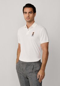 Biely polo s kontrastným svetlým golierom, s vyšitou postavou a logom "Hackett". Nosený s kockovanými nohavicami.