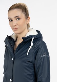 Giacca impermeabile blu navy con fodera in pelliccia sintetica bianca, cappuccio con coulisse e logo sulla spalla. Presenta bottoni a pressione e una texture liscia.