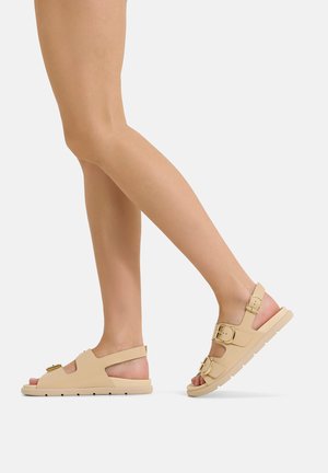 Person trägt beige offene Sandalen mit drei goldenen Schnallen, gezeigt beim Gehen vor einem einfachen hellen Hintergrund.