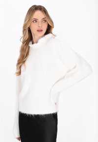 Witte gebreide trui met een pluizige kraag en manchetten, met een ontspannen pasvorm en textielstructuur. Gedragen over een zwarte, glanzende rok.