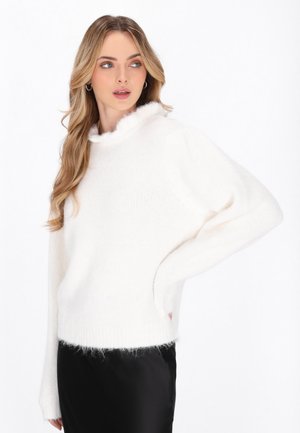 Witte gebreide trui met een pluizige kraag en manchetten, met een ontspannen pasvorm en textielstructuur. Gedragen over een zwarte, glanzende rok.