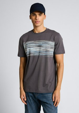 Mężczyzna w ciemnoszarej koszulce z wzorem poziomych pasków w kolorze jasnoniebieskim i białym na wysokości klatki piersiowej oraz czarnej czapce, stojący na tle jednolitego tła.