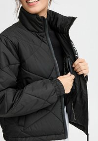 Giacca nera trapuntata con colletto alto e polsini elasticizzati. Presenta una tasca con zip all'interno, realizzata in materiale liscio e leggero.