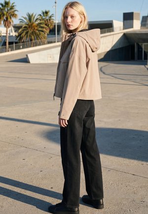 Junge Frau mit blonden Haaren, die eine beige Kapuzenjacke und schwarze, weite Hosen trägt, steht draußen bei Betonstrukturen und Palmen.