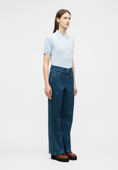 Γυναίκα όρθια, φοράει ανοιχτό μπλε πλεκτό μπλουζάκι με κοντά μανίκια και γιακά και ψηλόμεσα, φαρδιά μπλε τζιν με μικρά λευκά σχέδια, καφέ παπούτσια.