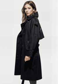 Mujer joven con cabello castaño claro vistiendo un largo abrigo negro, de pie de lado contra un fondo claro y simple.