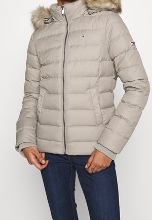 Personne portant une veste matelassée gris clair avec une capuche bordée de fourrure et un jean bleu, mains détendues près des poches.