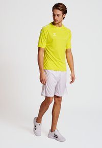 Geel sportshirt met korte mouwen, in combinatie met witte shorts en lichtgrijze sneakers met zwarte accenten.