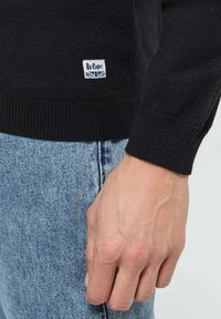 Pull en tricot noir avec des manches côtelées, associé à un jean bleu clair. Présente une petite étiquette avec "Le Coppe" et un design de Union Jack.