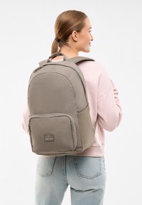 Rucsac gri cu o formă rotunjită, compartiment principal cu fermoar, buzunar frontal și bretele pentru umăr căptușite. Materialul pare durabil și texturat.