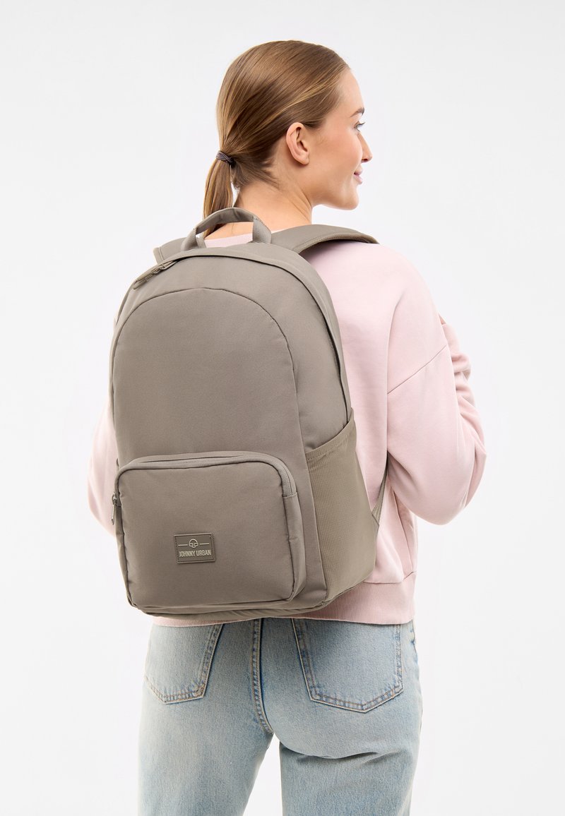 Rucsac gri cu o formă rotunjită, compartiment principal cu fermoar, buzunar frontal și bretele pentru umăr căptușite. Materialul pare durabil și texturat.