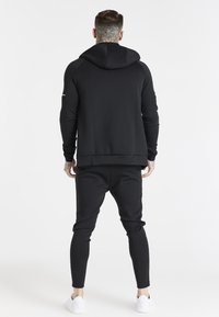 Sudadera negra con capucha y pantalones deportivos a juego hechos de tela suave. Presenta una capucha, puños acanalados y un diseño de ajuste slim. Combinado con zapatillas blancas.