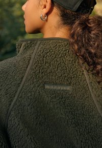 Olijfgroen fleece jack met een gestructureerde, pluche oppervlakte; heeft een zwart logo-label en contrasterende naden langs de randen.