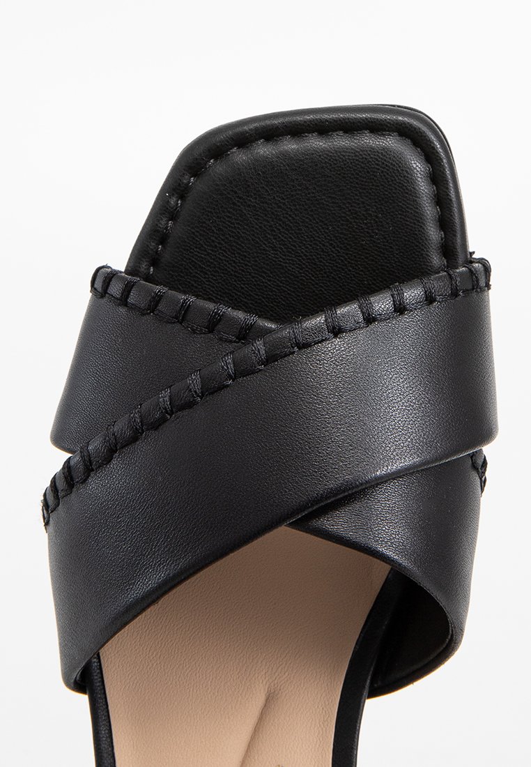 Sandalía negra de tacón alto con correas cruzadas, hecha de suave cuero sintético, que presenta un detalle trenzado a lo largo de los bordes y una punta cuadrada.