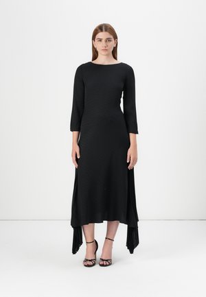 Model stands in a black, textured long-sleeve dress with a high-low hem, paired with black strappy heels, against a plain white background.