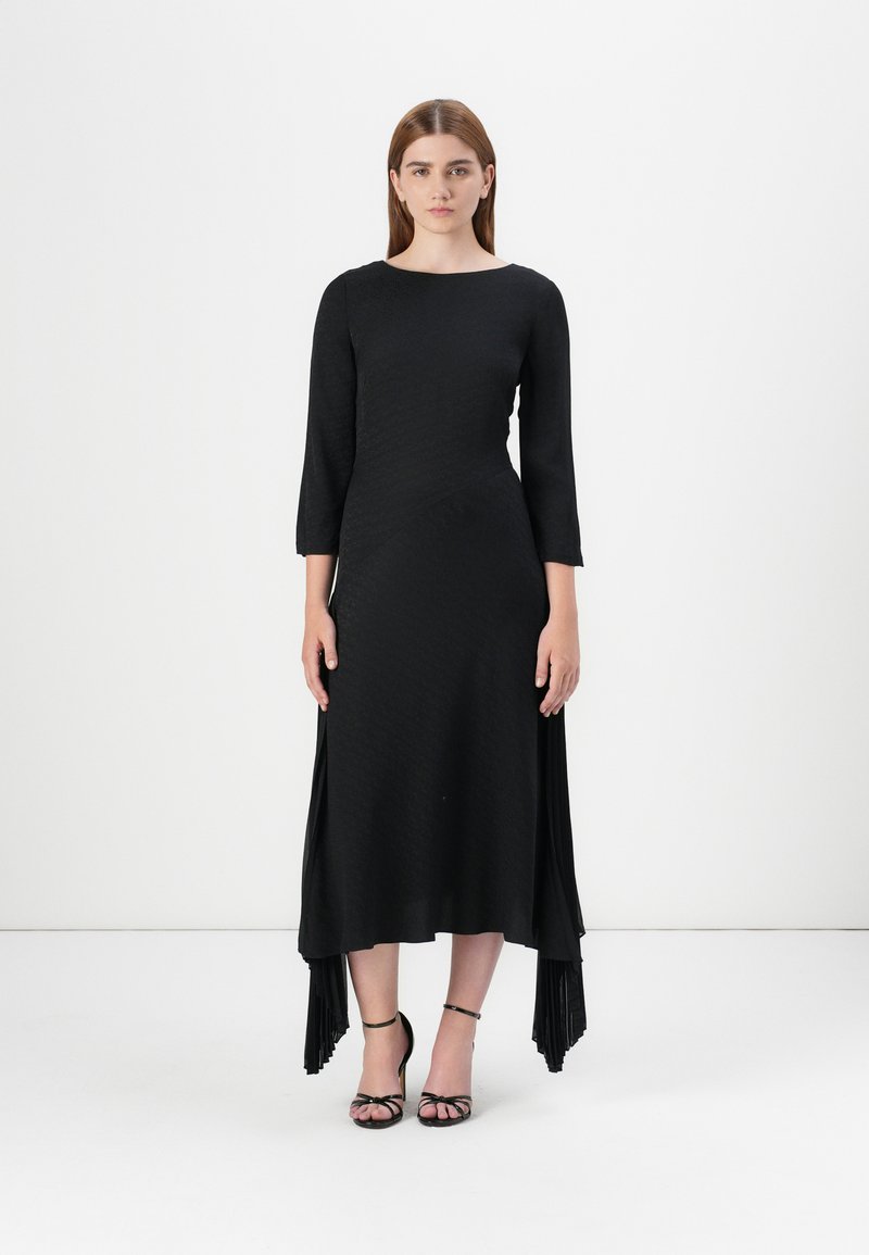 Model stands in a black, textured long-sleeve dress with a high-low hem, paired with black strappy heels, against a plain white background.
