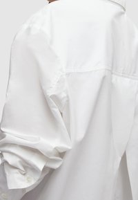 Chemise blanche avec une texture lisse et propre. Comprend des manches retroussées, des poignets boutonnés et un empiècement au dos. Style minimaliste.