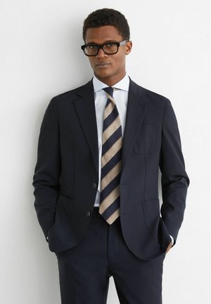 Muž v tmavomodrom obleku, bielej košeli, pásikatej béžovo-modrej kravate a čiernych okuliaroch, stojaci s rukami v vreckách oproti bielej stene.