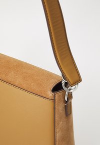 Sac à main en suède et cuir marron de forme rectangulaire, avec une bandoulière en cuir et des accents en métal argenté. Texture lisse sur le côté en cuir.