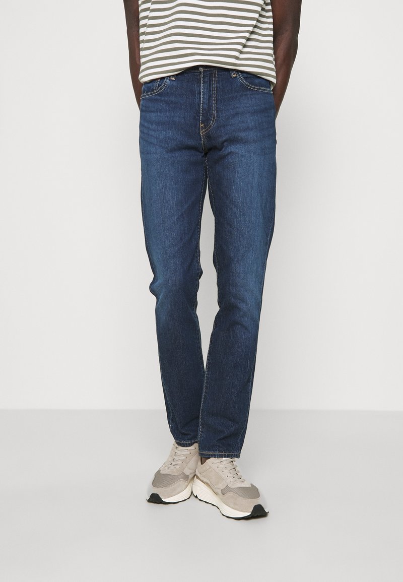 Dark blue denim jeans with a straight-leg cut and subtle stitching details. Worn with a striped shirt and light-colored sneakers.