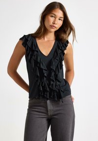 Top senza maniche nero con profondo scollo a V e balze che scendono lungo il davanti, abbinato a jeans grigi. Tessuto di texture liscia.