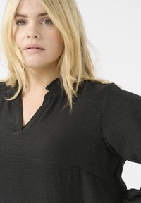 Blouse noire en tissu texturé, avec un décolleté en V et des détails subtils en forme de vagues. Coupe ample avec des manches trois-quarts.