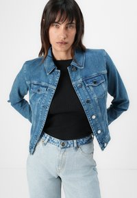 Denim jacket in light blue with a classic collar, front pockets, and silver buttons; paired with a black ribbed shirt and light blue jeans.