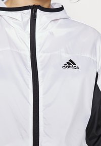 Bílá a černá zipová bunda z lesklé látky, s kapucí, černými akcenty a logem adidas na levé straně hrudi.