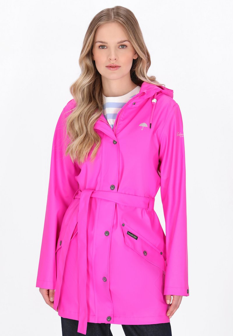 Femeie cu păr lung, ondulat, blond, purtând o haină de ploaie roz aprins cu curea peste o cămașă cu dungi deschise, cu fața spre înainte și o expresie neutră.