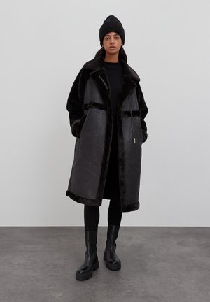Zwarte faux bonten jas met een brede kraag, ritssluiting aan de voorkant en zijzakken. Draag het over een zwart topje en leggins, gecombineerd met zwarte platformlaarzen.