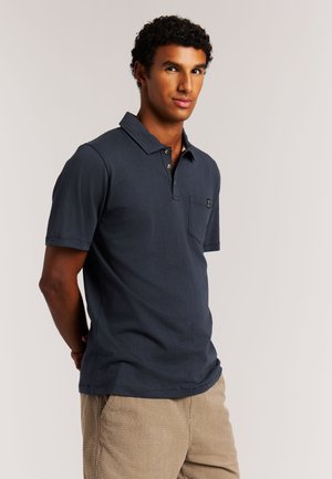 Homme aux cheveux courts et bouclés portant un polo bleu marine foncé et un pantalon beige texturé, debout devant un fond uni et clair.