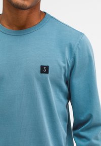 Langärmliges T-Shirt mit Rundhalsausschnitt aus weichem, hellblauem Baumwollstoff, mit einem kleinen schwarzen Quadrat-Logo, das einen weißen Buchstaben "J" auf der Brust zeigt.