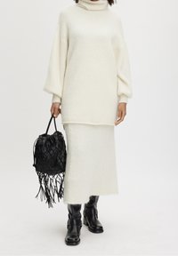 Sofá de punto blanco con un cuello alto, combinado con una falda larga a juego. Bolso de cubo negro tejido y botas negras hasta la rodilla.