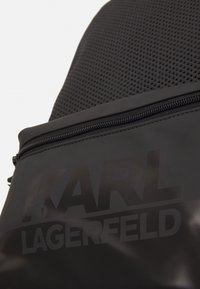 Czarny plecak z teksturowanym wierzchem, gładką dolną częścią, kieszenią na zamek błyskawiczny oraz wyraźnym logo "KARL LAGERFELD".