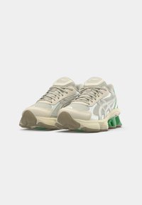 Beige athletic shoes with a mesh upper, reflective accents, and a green cushioned sole. Features lace closure and layered design.