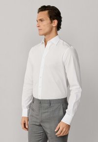 Mężczyzna ubrany w dopasowaną białą koszulę i szare, dopasowane spodnie, stojący na tle jednolitego jasnego tła, patrzący w bok.