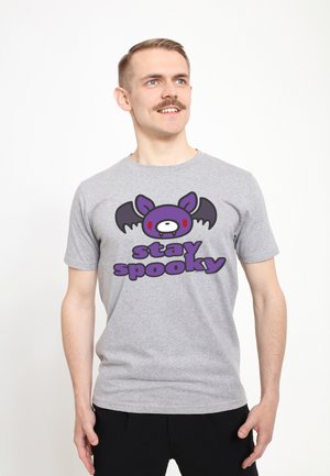 Uomo dalla pelle chiara con baffi che indossa una maglietta grigia con un disegno viola di un pipistrello e la scritta "stay spooky", in piedi su uno sfondo bianco.