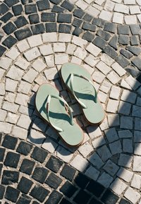 Paire de tongs vert clair reposant sur un pavement en pierre noir et blanc à motif circulaire sous la lumière du soleil.