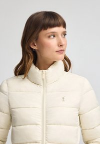 Krämfärgad puffjacka med hög krage och horisontell sömnad. Har en framficka med dragkedja och en liten logotyp på bröstet.