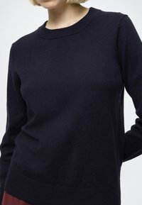 Tengerészkék kötött pulóver kerek nyakkivágással, hosszú ujjakkal, bordázott mandzsettákkal és szegéllyel. Puha tapintású, testhez simuló kialakítású, sima felületű.