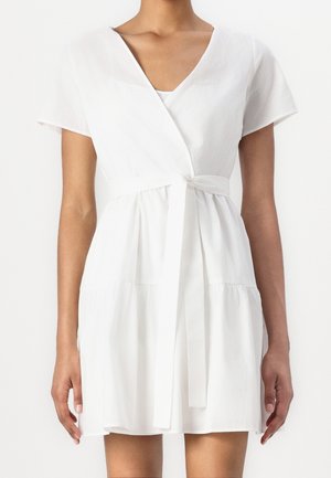 Personne mince portant une robe portefeuille blanche courte à manches courtes avec une ceinture nouée à la taille sur un fond uni.